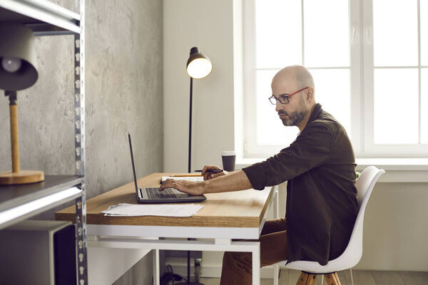 Focused serious adult business man working with documents and laptop in home office. Side view of casual man doing business remotely and checking reports while sitting in room with loft interior.