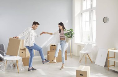 Happy young married couple dancing in bright room of their new home surrounded by cardboard boxes and minimal decor. Smiling woman holding hands with her husband, having fun together on moving day.