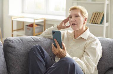 Mature woman sitting on gray sofa, holding smartphone, looking tired and frustrated during video call or phone conversation. Elderly lady touching her head, solving work tasks in living room at home.