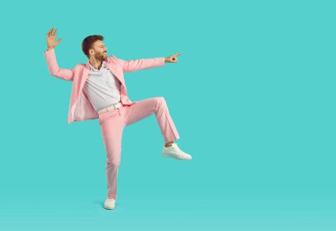Portrait of a young happy funny man wearing bright pink suit having fun and dancing isolated on a blue studio turquoise background and pointing index finger to the side to copy space.