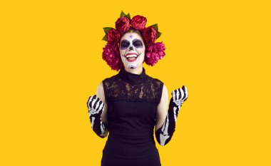 Positive emotions. Woman with sugar skull on Halloween rejoices in her success and victory, clenching her fists in joy. Woman with mexican face art and wreath on head, isolated on orange background.