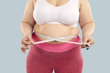 Cropped photo of faceless unrecognizable fat overweight woman wearing sportswear measure the waist with measuring tape isolated on studio blue background. Obesity, weight control concept.