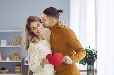 Young couple in love enjoying Valentines Day at home. Happy people hugging and kissing on a romantic date at home. Loving man embracing his woman, kissing her and whispering words of love