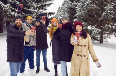 Cheerful friends during winter walk raise up paper cups with hot drink cheering and greeting you. Happy people in warm clothes together cheerfully shouting looking into camera holding disposable cups.