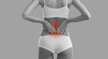 Woman suffers from pain and touches lower back. BW backside view from behind lady with slim figure standing on grey background holding hands on red highlighted pain area. Spine problem, injury concept