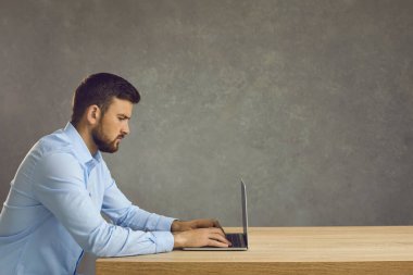 Serious focused smart programmer or employee working with laptop on deadline project. Side view of a young caucasian man sitting at a table on a gray background with free space for text.