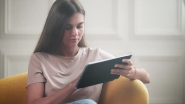 Tablet bilgisayarı olan uzun saçlı kız. Genç bir kadın evinde koltukta dinleniyor ve sosyal medyaya veya internetten alışveriş yapıyor.