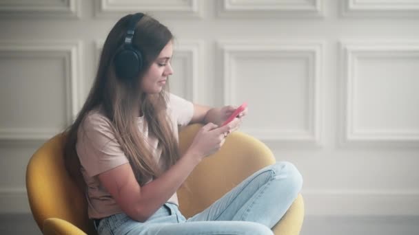 十几岁的女孩使用智能手机 用耳机听音乐 一个人坐在家里 欣赏最喜爱的歌曲 周末在隔离流行时期消磨时间 快乐的年轻女生 — 图库视频影像