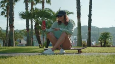 Sportif kızlar kaykayda oturur ve parkta avuçlarıyla akıllı telefondan görüntülü sohbet ederler. Neşeli genç bir kadın selam vermek için el sallıyor ya da elveda demek için.