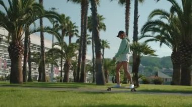 Sportif bir kızın kaykayla eğlendiği yavaş çekim. Palmiyeler arasında uzun tahta sürüşü ile aktif boş zaman