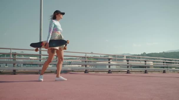 年轻的女人看起来很有运动精神 在城市里散步 还带着一个长板 快乐而无忧无虑的女孩在滑板 慢动作后享受时光 — 图库视频影像
