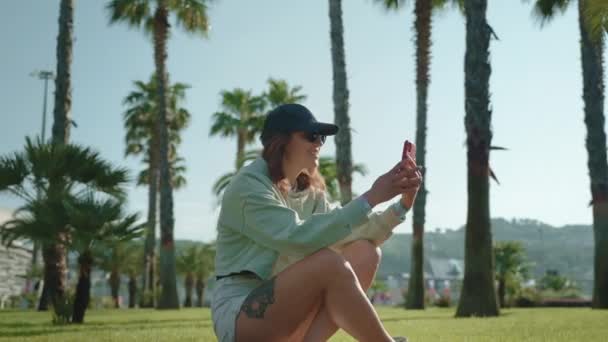 在公园的长板上坐着一个看起来很有运动精神的小女孩 她用智能手机打了一个视频电话 在明亮的阳光照射下的手掌间看 — 图库视频影像