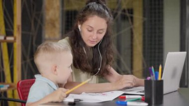 Genç anne, bebek bakıcısı ve küçük çocuk evde vakit geçiriyorlar. Kadın dizüstü bilgisayar ve kulaklık kullanıyor. Oğlu oynuyor.