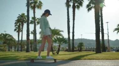 Güneşli bir günde uzun kaykayla gezen güzel genç bir kadın palmiye ağaçlarının yanından geçiyor, güzel bir tropikal şehirde yavaş çekim yapıyor, yaz tatilinde spor hobisi ve eğlence, gençlik alt kültürü ve spor yapıyor.