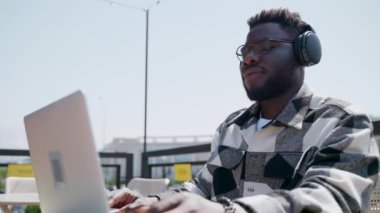 Genç Afro-Amerikan bir adam açık şehir kafesinde dinleniyor. En sevdiği müziği kablosuz kulaklıkla dinleyip dizüstü bilgisayar kullanıyor.