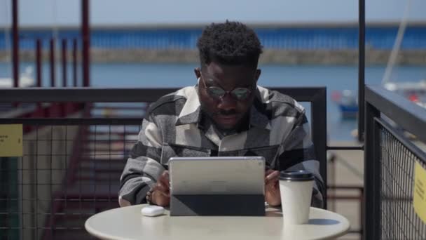 アフリカ系アメリカ人の男性は海の近くの屋外カフェで一日を過ごす 彼はネットサーフィンやソーシャルメディアをデジタルタブレットで閲覧 — ストック動画