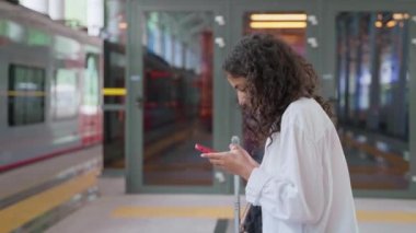 Platformda bavullu genç bir esmer. İnternette sörf yapmak için telefon kullanan ya da gecikmiş treni beklerken sohbet eden kadın yolcu.