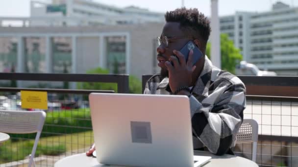 ハンサムな黒人男性がノートパソコンで働いてるカフェで携帯電話で話してる肖像画アフリカ系アメリカ人の男が遠隔操作で働いてるビジネスオーナーが携帯電話で電話を呼んでる — ストック動画
