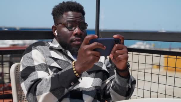 Jovem Negro Com Smartphone Descansando Terraço Perto Mar Dia Ensolarado — Vídeo de Stock