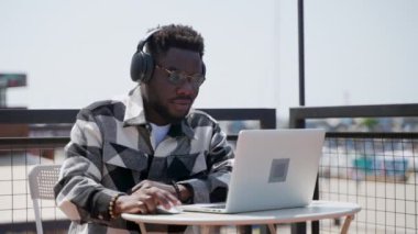 Afro-Amerikan öğrenci kafede dizüstü bilgisayar kullanıyor, siyahi adam sosyal medyada içeriği izliyor, kulaklıkla müzik dinliyor, sokak kafesinde portre çekiyor.