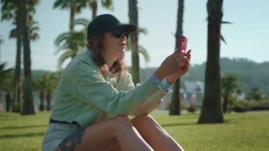 Spor kıyafetli bir kız parkta çimlerde avuç içiyle oturuyor ve cep telefonuyla video görüşmesi yapıyor. Dışarıda cep telefonuyla vakit geçirmek.