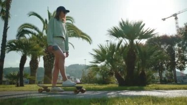 Genç bir kadın aktif yaşam tarzını tercih ediyor ve yeşil güneşli parkta palmiyeli bir kaykay gezisine çıkacak. Kadın ayağı kaykayı hareket ettiriyor.