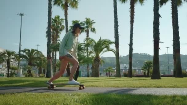 緑のヤシの木の間の公園のパスに沿ってスケートボードを楽しむスポーティガール スローモーション アクティブな趣味と屋外レジャー — ストック動画