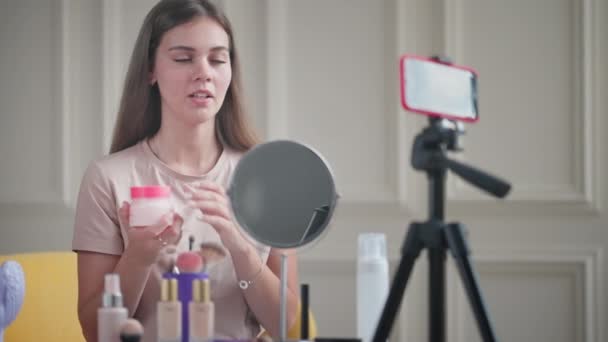 年轻女人在她的美容博客上提供有关皮肤护理和化妆的指导 她闻到了奶油的味道并解释了如何使用它 在三脚架上记录视讯和智能手机 — 图库视频影像