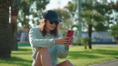 Modern mobil cihazlı havalı bir genç kız güneşli bir yaz gününde parkta portre açık havada, akıllı telefon kullanan genç bir kadın sosyal medyada paylaşımlar, yaz tatilinde tembellik.