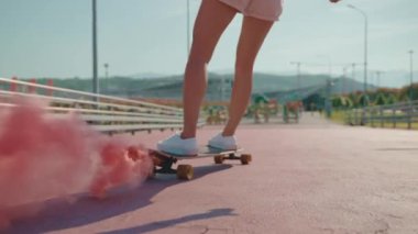 Pembe renkli sis bombası, yakın çekim görüntüsü olan uzun panolu genç kız kaykayla kayıyor, eğleniyor ve eğleniyor, spor ve eğlence için spor ayakkabıları ve spor hobileri var.