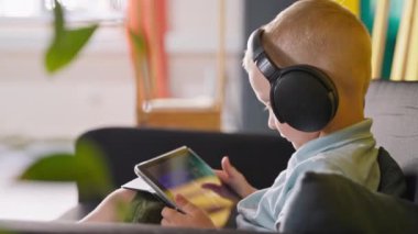 Aletlerle evde vakit geçirmek. Küçük çocuk kanepede oturup dijital tablet ve kablosuz kulaklık kullanarak çizgi film izliyor.