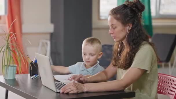ノートパソコンで働くか オンライン訓練を取るイヤフォンの女性 彼女の小さな子供は絵を描くが 母親が何をするかについて — ストック動画