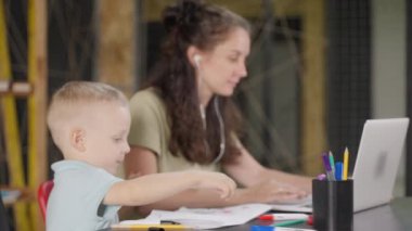 Küçük çocuk, annesi uzaktan kumandalı işleriyle meşgulken resim yapmaya zaman harcıyor. Çocuğa odaklan, arka planda dizüstü bilgisayarı olan kadına. Merkez ofis ve aile.