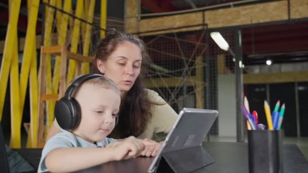 小さな男の子はデジタルタブレットで学習ゲームをプレイ 母親は彼の行動を見てチェックする 電子工学による教育活動 — ストック動画