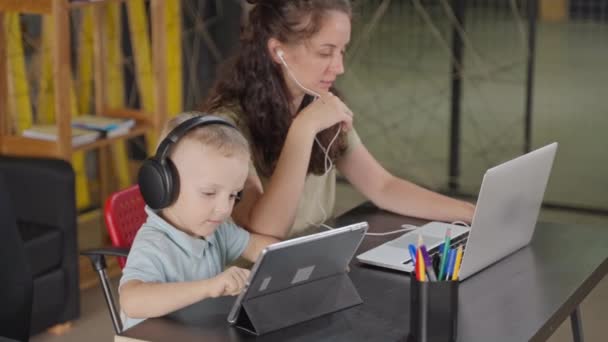 自宅のオフィスでリモートで働く女性 彼女はラップトップでビデオ会議をしているとき ヘッドフォンの彼女の小さな息子はタブレットコンピュータ上のゲームに従事した キャリアと家族 — ストック動画