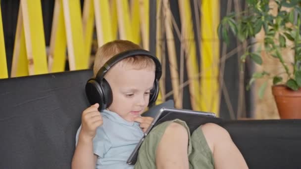 坐在沙发上 坐在平板电脑上看卡通片或录像的大无线耳机里 蹒跚学步的男孩 带小玩意的室内休闲活动 — 图库视频影像