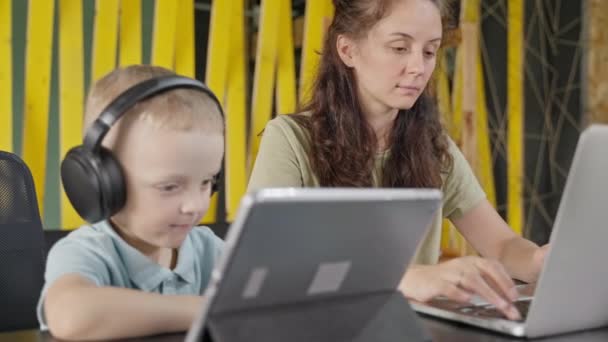 家で働く女性 ヘッドフォンで彼女の小さな息子がデジタルタブレット上の漫画や教育ビデオを見ているときにラップトップを使用してフリーランスの母親 キャリアと家族 — ストック動画