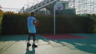 Güneşli bir sabahta Afrikalı Amerikalı erkek basketbol sahasında antrenman yapıyor. Topu sektiriyor ve atışa hazırlanıyor.