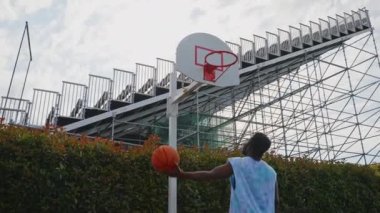 Açık havada basketbol antrenmanı yapan Afrikalı bir Amerikalı yavaş çekim yapıyor. Adam tek elle ateş ediyor ama potayı ıskalıyor.