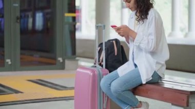 Bagajlı kadın tren istasyonunda bankta oturuyor, akıllı telefondan zaman çizelgesini kontrol ediyor, terminalde ücretsiz kablosuz internet kullanıyor, trenle seyahat ediyor, şehir sakinleri ve banliyö sakinleri için toplu taşıma araçları kullanıyor.