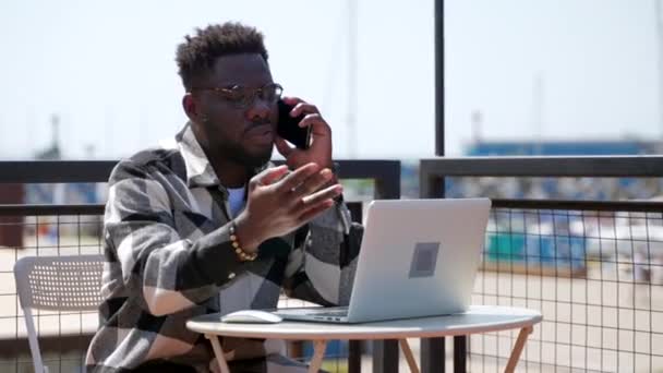 電話で話している黒人の遅い動きのショットといくつかのビジネス上の問題を解決するためにラップトップを使用しています 屋外のシティカフェで働く忙しい人 — ストック動画