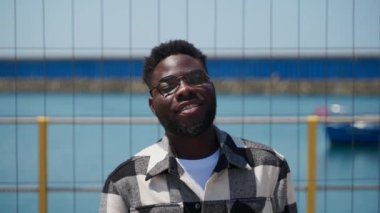 Gözlüklü ve siyah beyaz kareli gömlekli neşeli bir Afro-Amerikalı 'nın orta pozu. Bulanık denize karşı duruyor metal tel çitlerin arkasında kayıkla