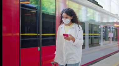 Akıllı telefonu ve yüzünde koruyucu maskesi olan kadın yolcu tren platformunda yürüyor, arabasına biniyor, covid-19 salgını, virüs salgını mevsiminde banliyö demiryollarında seyahat ediyor.