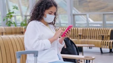 Covid-19 enfeksiyonunun önlenmesi için önleme tedbirleri, havaalanı veya tren istasyonunun kalkış salonunda maskeli yolcu, kadın cep telefonuyla internette sörf ve biniş bekliyor