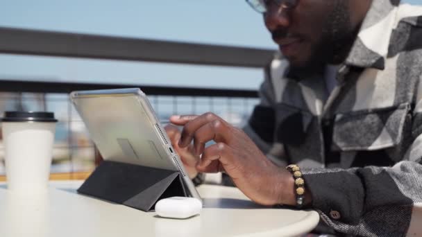 デジタルタブレットを使用して黒人男性のクローズアップショットは 仕事や自由な時間を過ごすために 彼は屋外のカフェのテーブルに座って 近くのコーヒーカップ — ストック動画