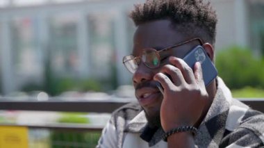 Gözlüklü genç siyahi adam dışarıda telefonla konuşuyor, yakın plan. Bulanık şehir görüşüne karşı konuşan kişi
