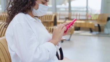 Terminalde uçuş ya da tren beklerken akıllı telefondan internette sörf yapan kadın, Covid-19 virüs enfeksiyonundan korunmak için yüz maskesi takıyor, salgın sırasında uçak ya da demiryolu ile seyahat ediyor.