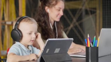 Kulaklıklı mutlu küçük çocuk internetten çizgi film ve video izliyor, annesiyle evde oturuyor, yetişkinler ve çocuklar için modern aletler, sosyal medya bağımlılığı ve çevrimiçi servis