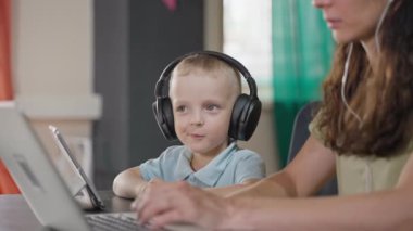 Kablosuz kulaklıklı çocuk, annesi evde dizüstü bilgisayarla, çocuklar ve yetişkinler için modern aletlerle, internet bağımlılığı ve boş zamanlarında tablet kullanıyor.