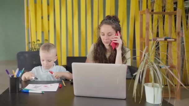 産前産後の休暇中の母親は自宅から遠隔地で働いています小さな息子と一緒に座って携帯電話で電話をかけインターネットでノートパソコンを使って仕事をしています — ストック動画